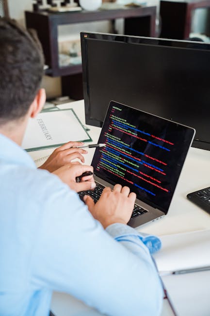 A person working at a desk with a laptop displaying lines of colorful code on the screen. The individual is wearing a light blue shirt and is using a wireless mouse with their right hand while their l