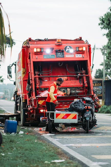 A worker in a red and yellow uniform is operating a large red rubbish collection truck on a paved urban street, positioned with its rear open for waste disposal. The truck's rear compartment reveals m